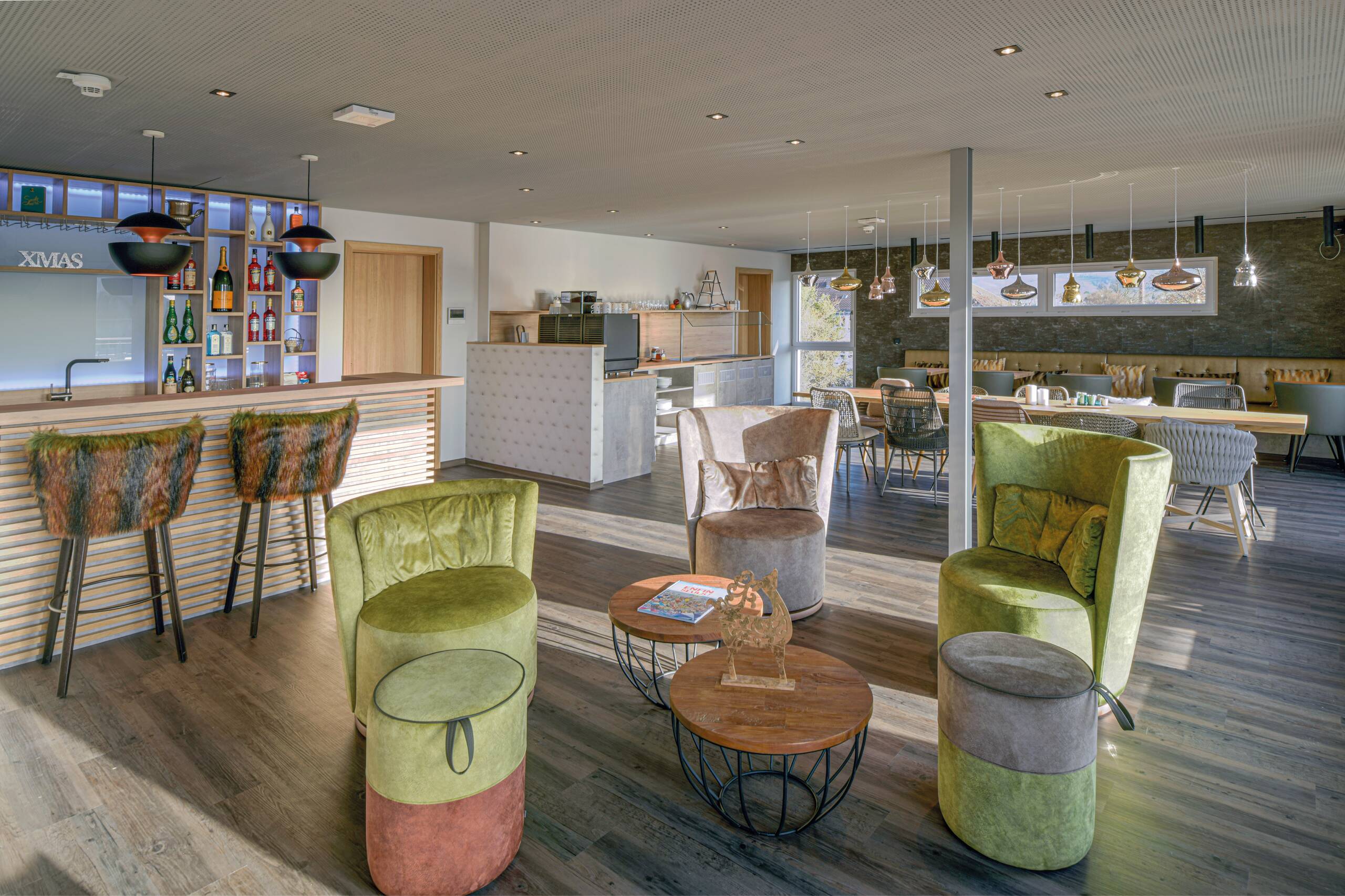 Aparthotel in Holzbauweise Eine moderne, helle Lounge mit einer Bar im Hintergrund. Es gibt gemütliche Sitzgelegenheiten, darunter zwei grüne Sessel, eine grau-beige Couch und runde Holztische. Hinter der Bar sind Regale mit verschiedenen Getränken. Das Ambiente ist einladend und stylisch.