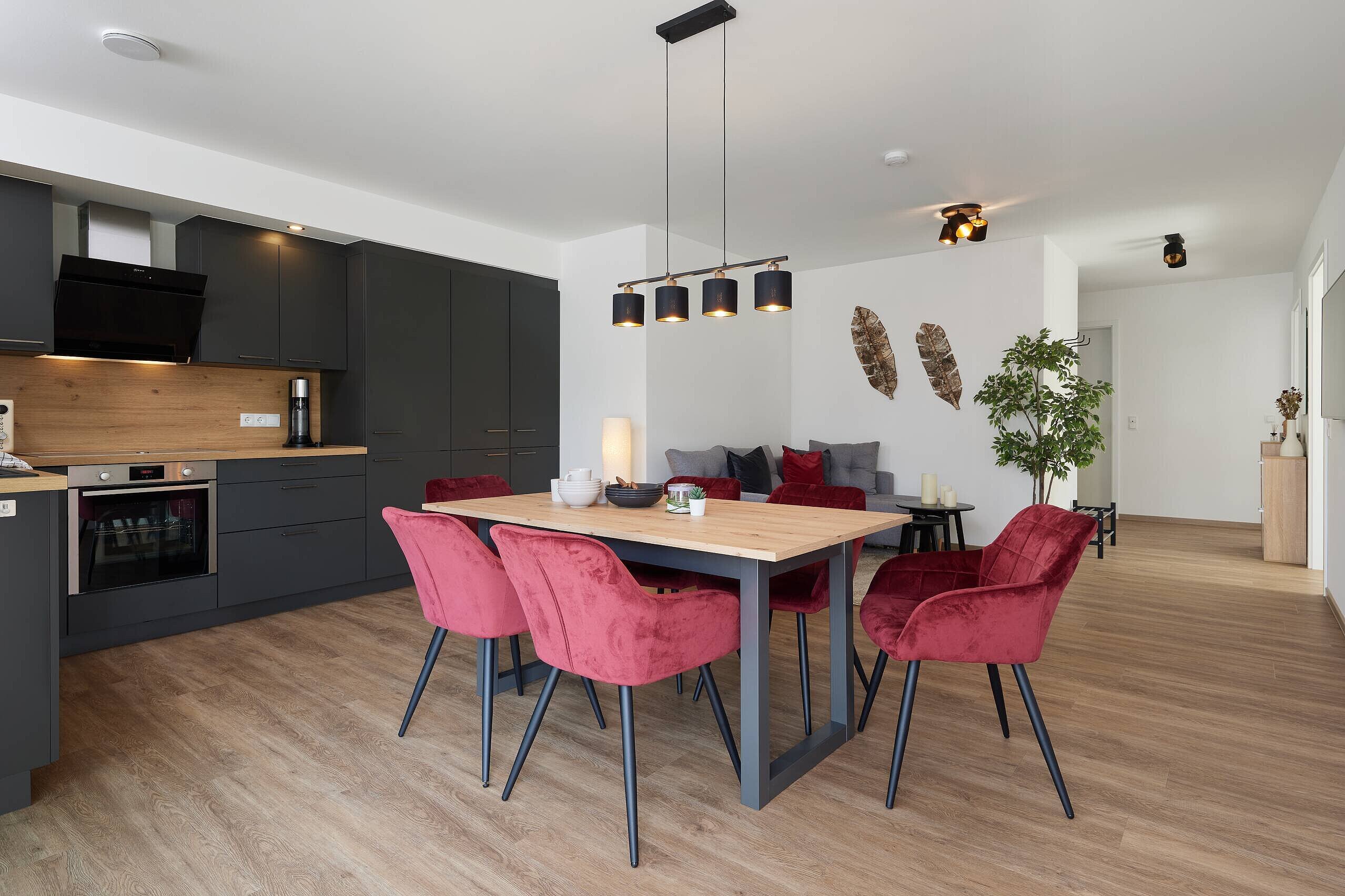 Offener Koch- und Essbereich Ein modern eingerichteter Wohnbereich mit einer offenen Küche. Am Tisch mit einer Holzplatte stehen sechs rote Stühle. Im Hintergrund sind ein Sofa mit Kissen und eine Pflanze zu sehen. Die Beleuchtung hängt über dem Tisch und schafft eine gemütliche Atmosphäre.