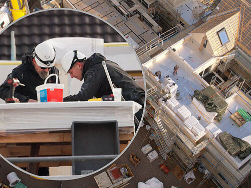 Zwei Bauarbeiter in Schutzhelmen arbeiten gemeinsam an einer Baustelle. Im Hintergrund sieht man ein unvollendetes Gebäude mit Gerüst und Bauteilen. Die Arbeiter sind auf einem Tisch mit Werkzeug und Materialien beschäftigt.