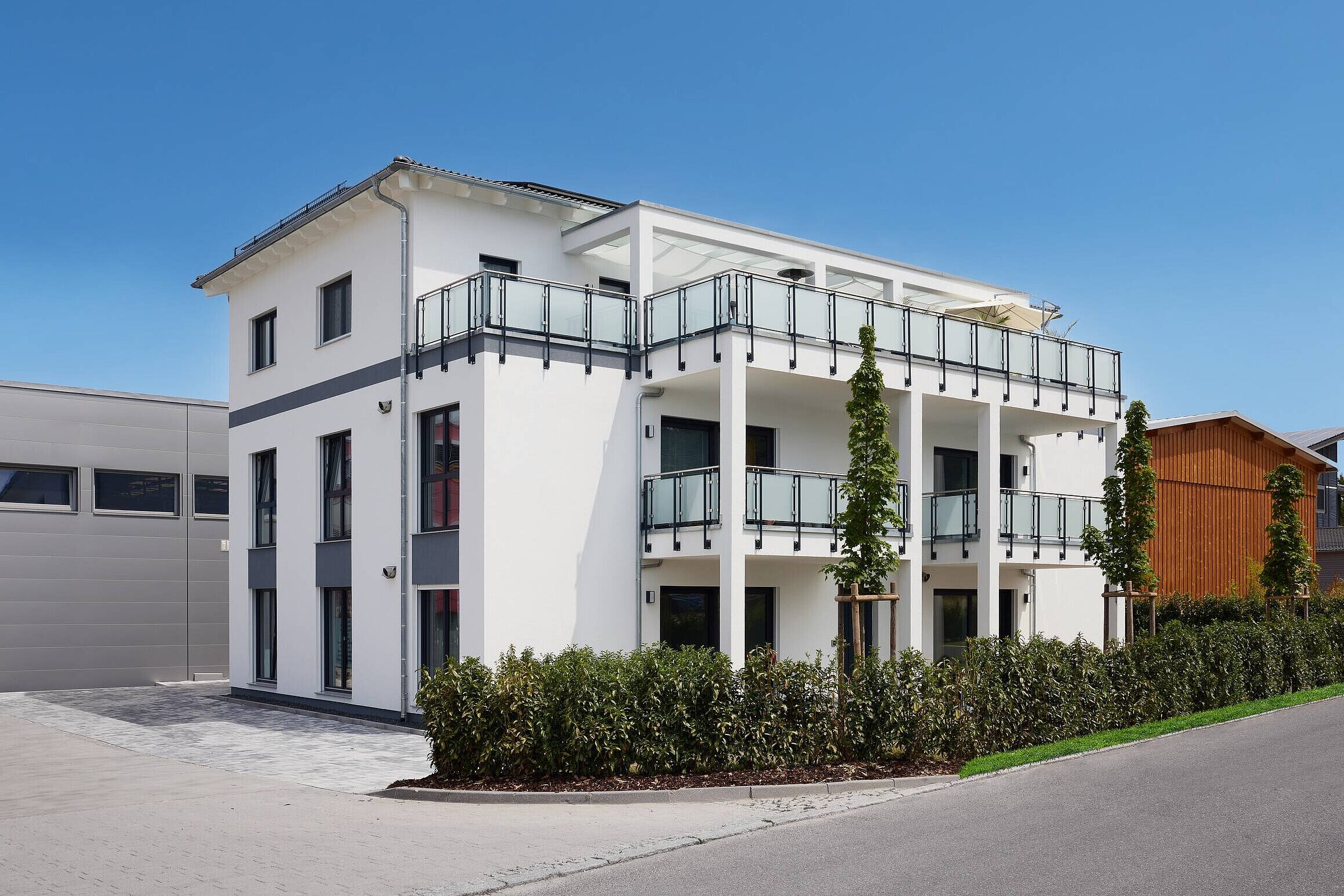 dreigeschossiges Mehrfamilienhaus Ein modernes, dreigeschossiges Wohnhaus in Weiß mit Balkonen aus Glas. Die Fassade hat große Fenster und graue Akzente. Im Vordergrund eine gepflasterte Zufahrt und grünes Gestrüpp. Im Hintergrund ist ein graues und ein holzverkleidetes Gebäude sichtbar. Der Himmel ist blau und klar.