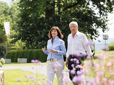 Heidi Weber-Mühleck und Hans Weber gehen zusammen durch den Erlenpark. Im Hintergrund sind grüne Bäume und bunte Blumen sichtbar. Die Atmosphäre wirkt freundlich und einladend.