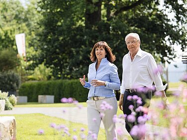 Heidi Weber-Mühleck und Hans Weber gehen zusammen durch den Erlenpark. Im Hintergrund sind grüne Bäume und bunte Blumen sichtbar. Die Atmosphäre wirkt freundlich und einladend.