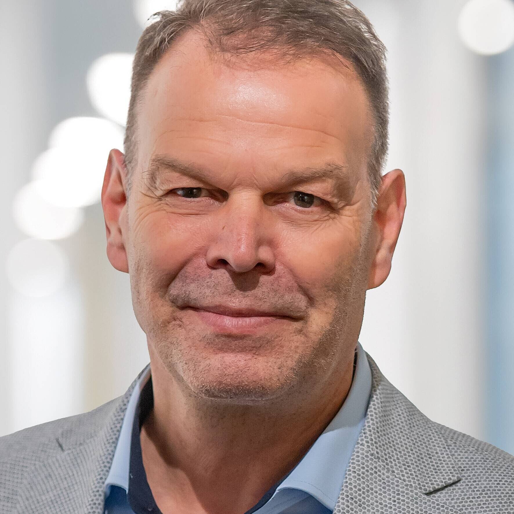 Porträt eines Mannes mittleren Alters mit kurzem, grauem Haar. Er trägt einen hellgrauen Blazer und ein hellblaues Hemd. Der Hintergrund ist unscharf und hell, mit weichen Lichtpunkten. Der Mann hat einen freundlichen Gesichtsausdruck und schaut direkt in die Kamera.