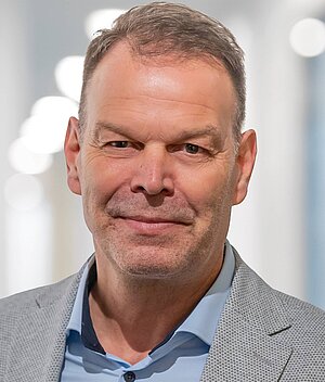 Porträt eines Mannes mittleren Alters mit kurzem, grauem Haar. Er trägt einen hellgrauen Blazer und ein hellblaues Hemd. Der Hintergrund ist unscharf und hell, mit weichen Lichtpunkten. Der Mann hat einen freundlichen Gesichtsausdruck und schaut direkt in die Kamera.