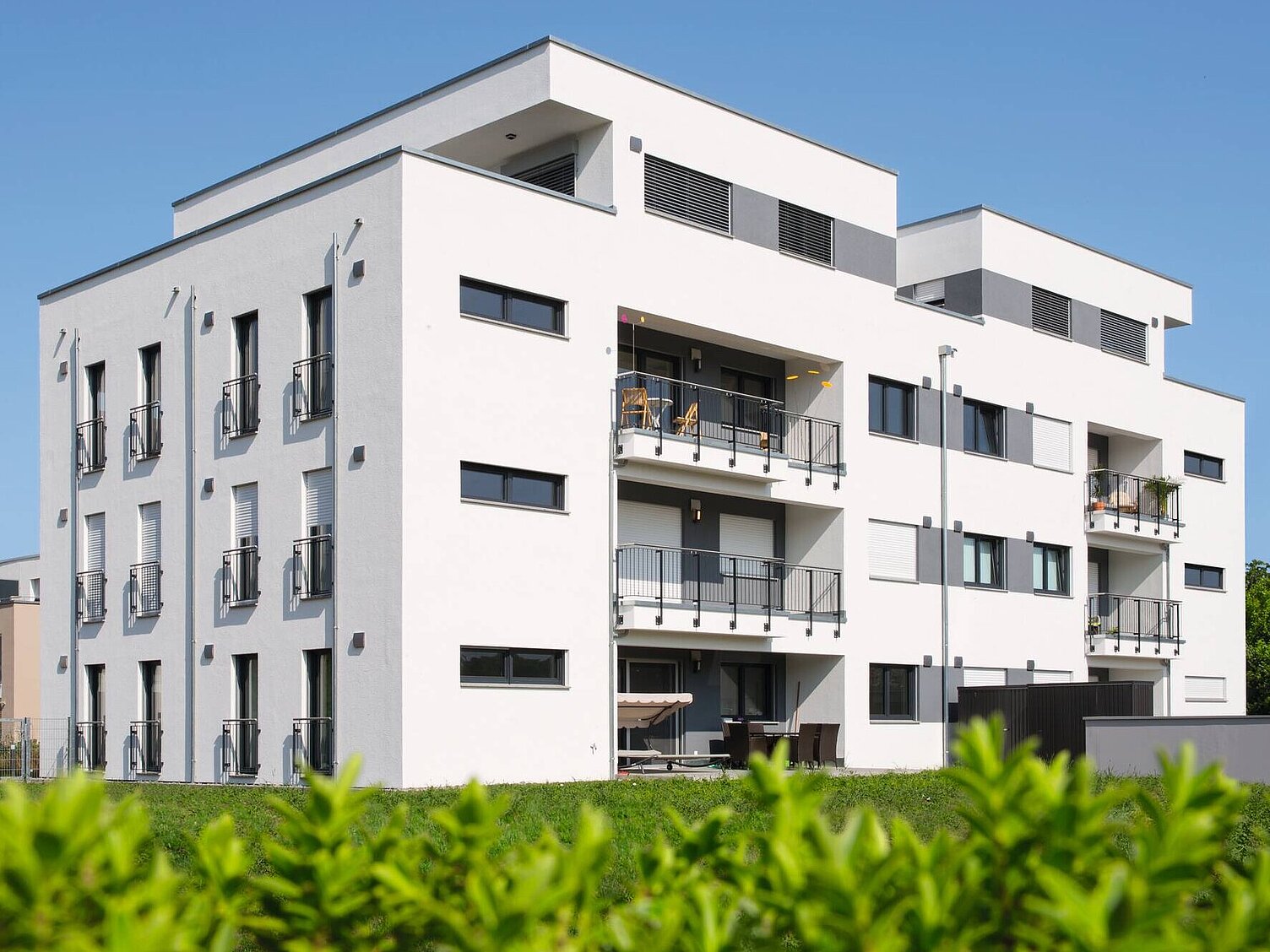 Ein modernes Wohngebäude mit mehreren Etagen ist im Vordergrund zu sehen. Die Fassade ist weiß, mit großen Fenstern und Balkonen. Im Hintergrund sind weitere Gebäude und ein grüner Bereich zu erkennen. Der Himmel ist klar und blau.