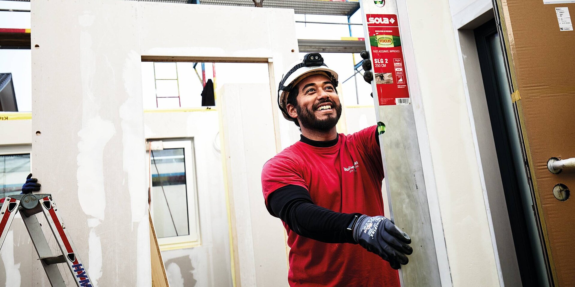 Ein lächelnder Bauarbeiter in einer roten T-Shirt und Handschuhen hält ein Messgerät. Er steht auf einer Baustelle mit Wänden und einem Fenster im Hintergrund. Eine Leiter ist sichtbar, und im Hintergrund hängen Baugerüste.