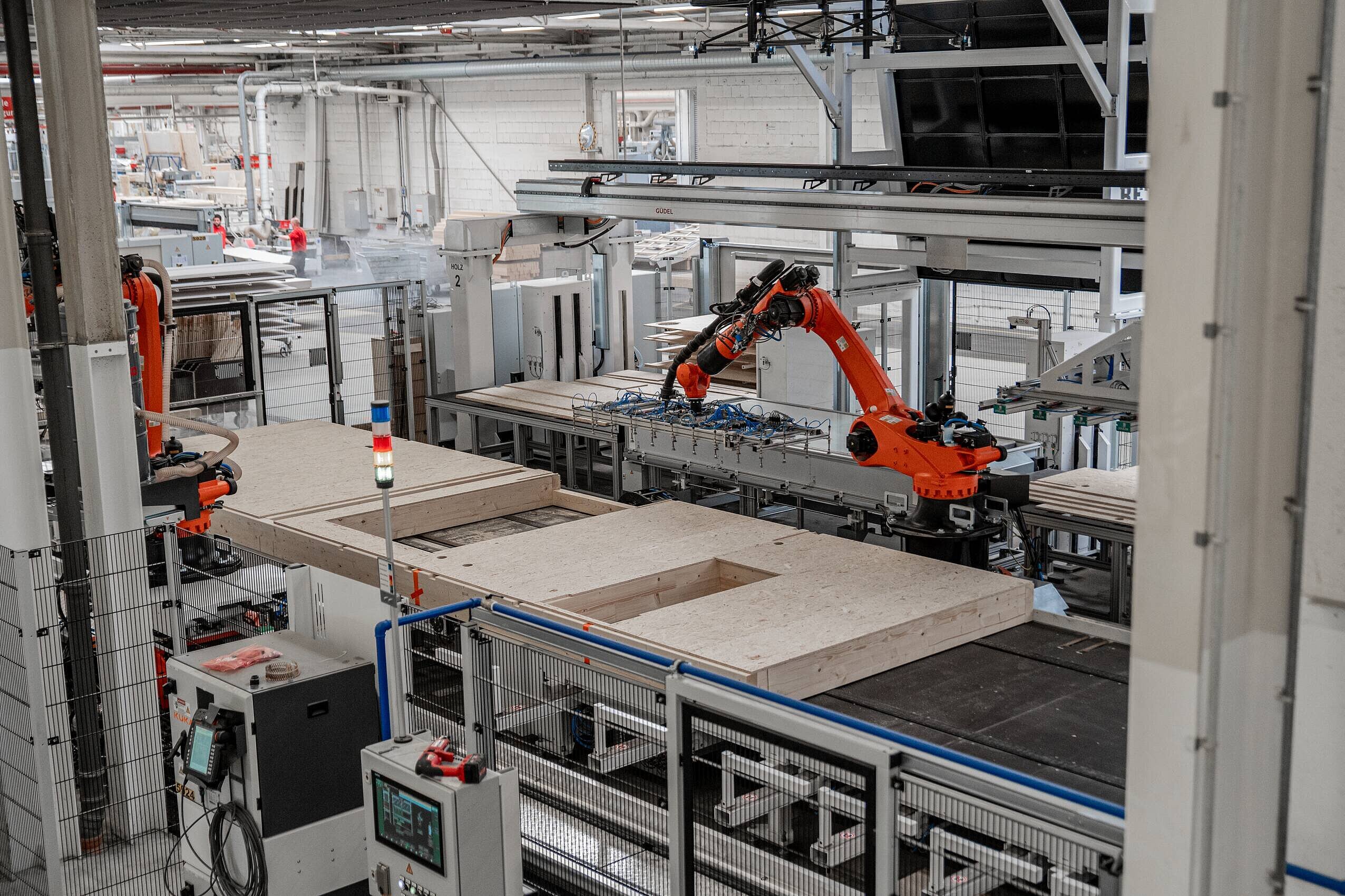 Robotik im Hausbau In einem modernen Produktionsraum ist ein roter Roboterarm zu sehen, der Innenwände produziert. Die Wände liegen auf einem Förderband, das für die Montage oder Bearbeitung vorgesehen ist. Im Hintergrund sind weitere Maschinen und Werkzeuge sichtbar.