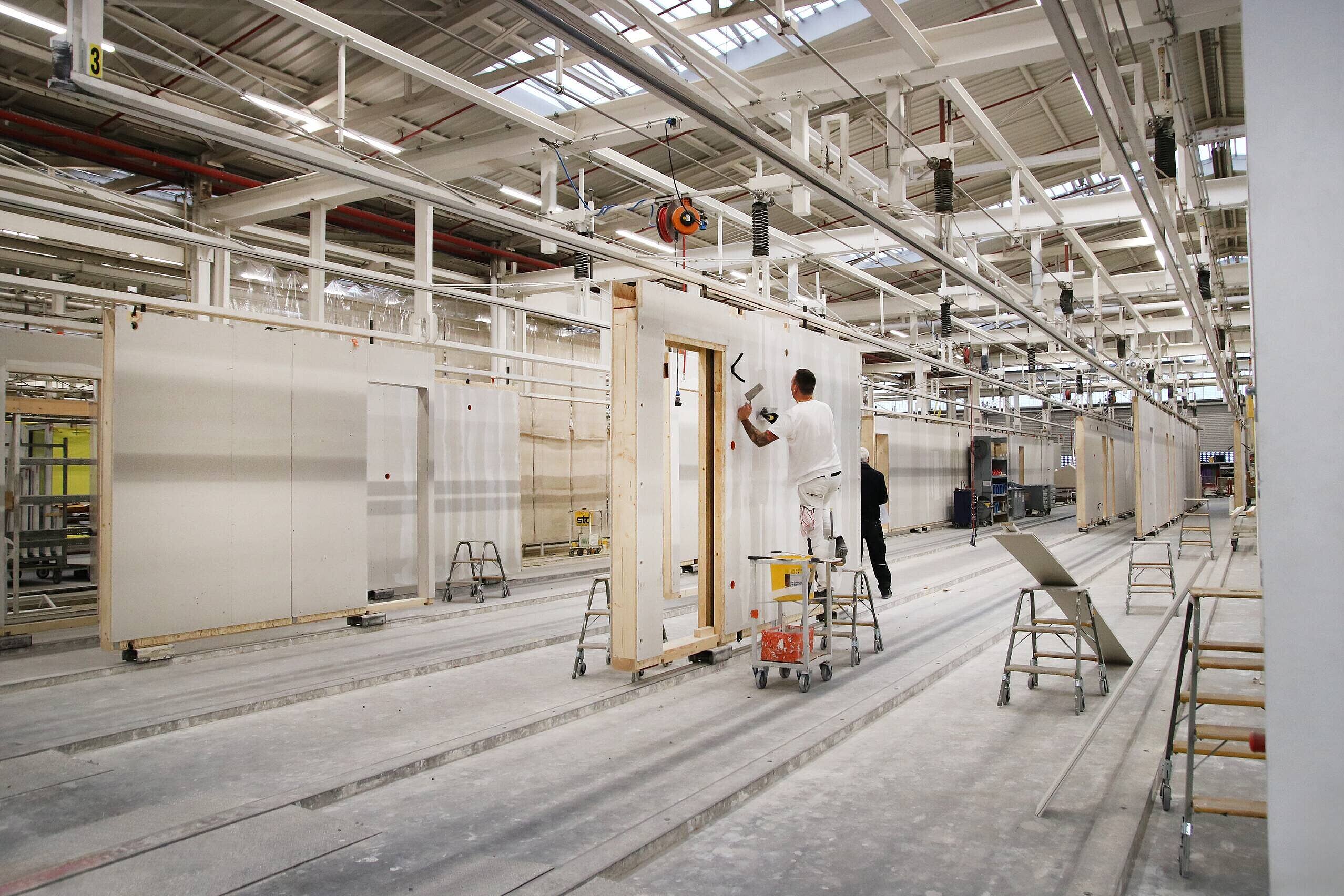 In einer Werkhalle sind mehrere Baustellen mit Holzrahmen zu sehen. Zwei Personen arbeiten an den Wänden, eine steht auf einer Leiter und streicht die Wand mit einer Bürste. Der Boden ist staubig, und es sind Baugeräte und Materialien sichtbar.