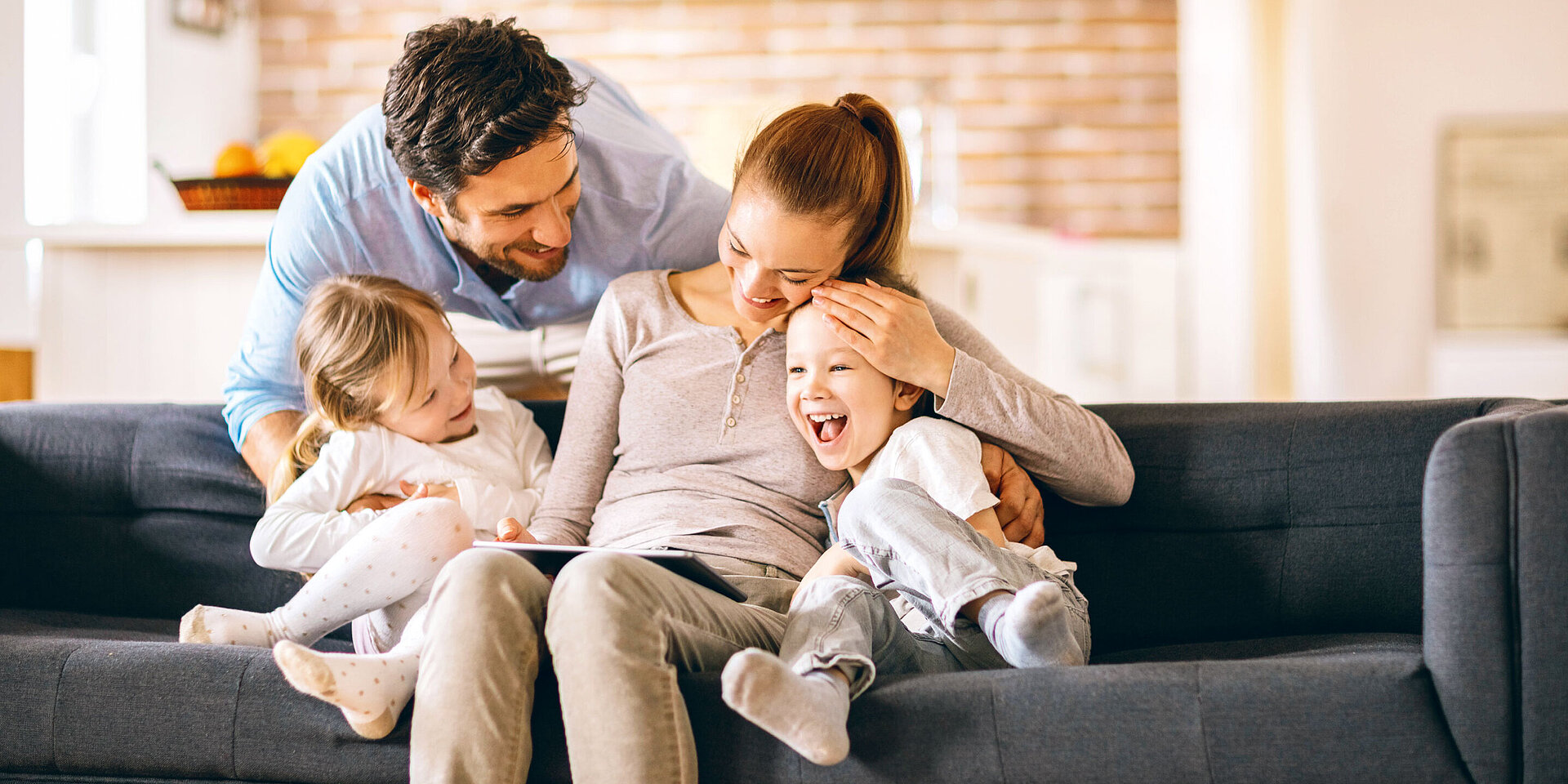 Eine Familie sitzt glücklich auf einem Sofa. Der Vater beugt sich über ein Kind, während die Mutter das andere Kind umarmt. Alle lachen und zeigen Freude, was eine liebevolle Atmosphäre in ihrem Zuhause vermittelt.