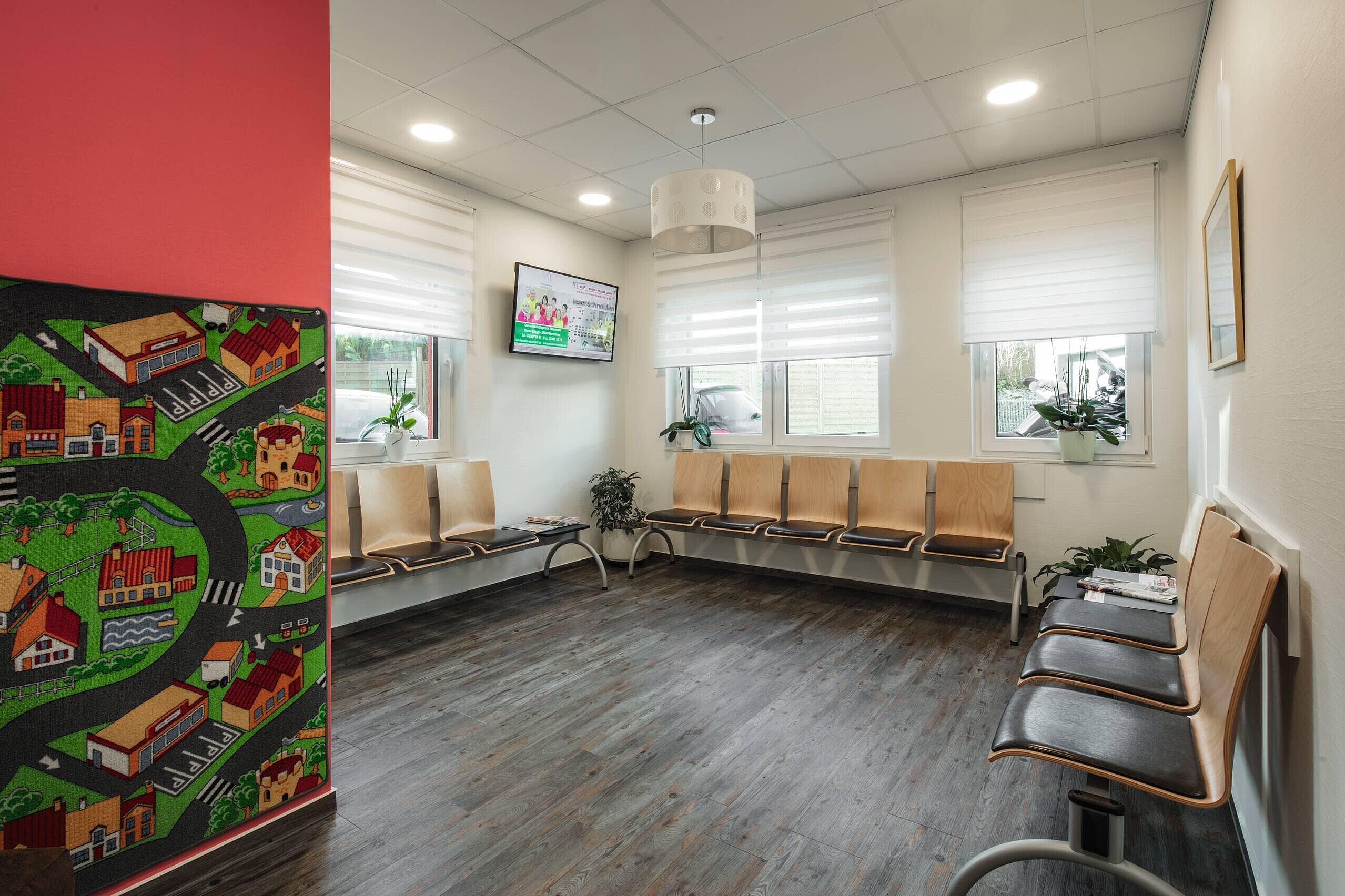 Ein Wartezimmer mit Holzbankreihen an den Wänden. An einem Fenster stehen Pflanzen und es gibt einen Fernseher, der Informationen anzeigt. Der Boden ist aus Holz, und die Wände sind in sanften Farben gestaltet.