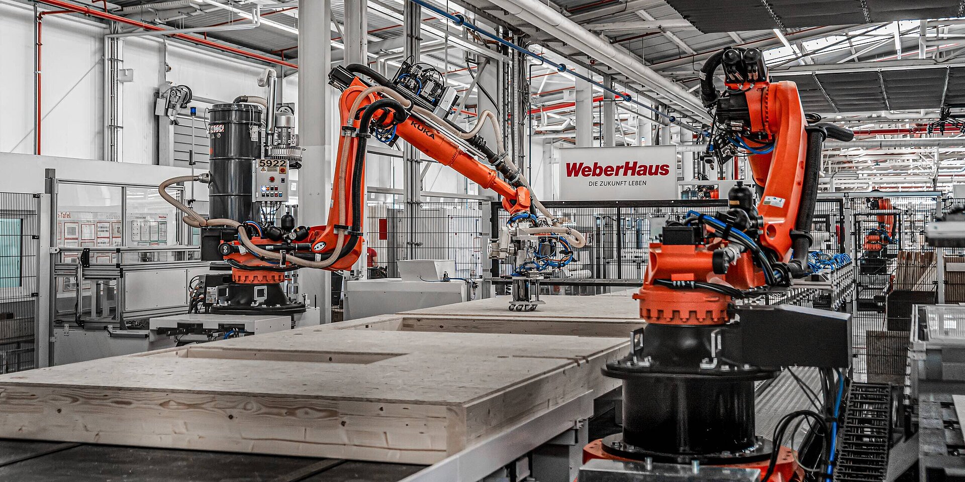 In einer modernen Fabrikhalle von WeberHaus arbeiten zwei orangefarbene Industrieroboter an einer Holzplatte. Im Hintergrund ist ein Schild mit der Aufschrift „WeberHaus – Die Zukunft leben“ zu sehen. 