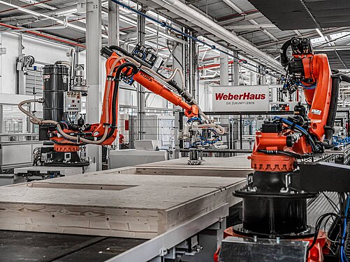 In einer modernen Fabrikhalle von WeberHaus arbeiten zwei orangefarbene Industrieroboter an einer Holzplatte. Im Hintergrund ist ein Schild mit der Aufschrift „WeberHaus – Die Zukunft leben“ zu sehen. 