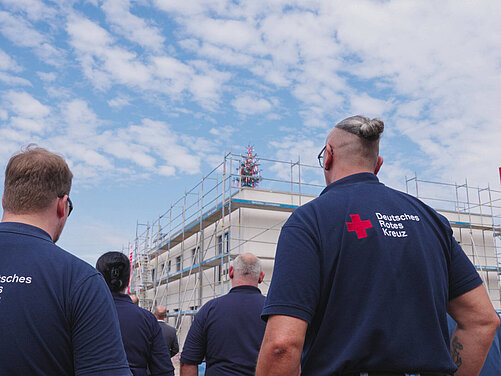 Gruppe von Mitarbeitern des Deutschen Roten Kreuzes, die auf ein Bauprojekt schauen. Im Hintergrund ist ein Gerüst zu sehen, das einen unvollendeten Gebäudeteil umgibt. Der Himmel ist blau mit einigen Wolken.