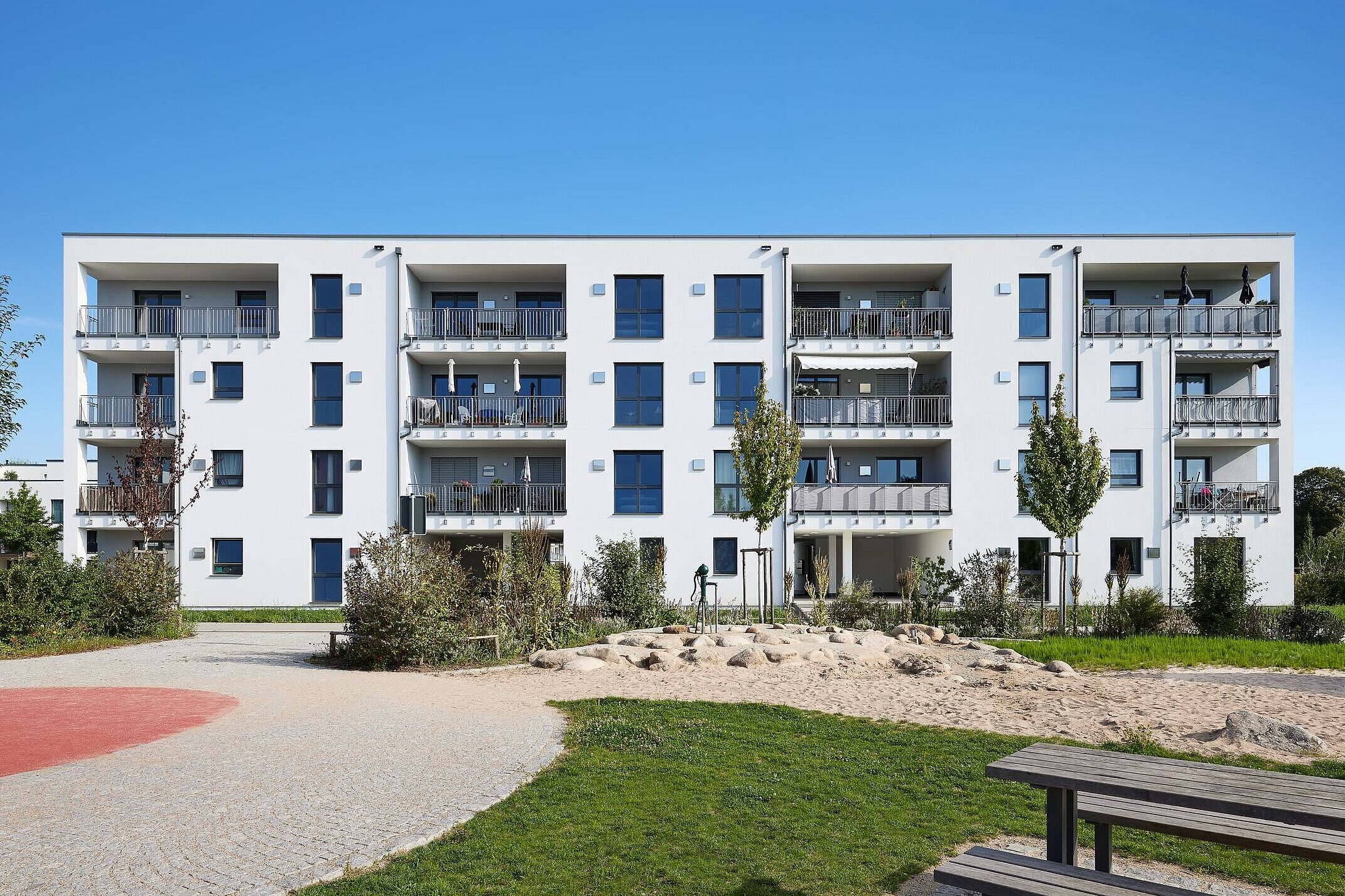 Mehrfamilienhaus in ökologischer Holzbauweise Moderner Wohnbau mit drei Etagen, großen Fenstern und Balkonen. Umgeben von grünen Flächen, Bäumen und einem Spielplatz mit Sandbereich. Der Himmel ist klar und blau.