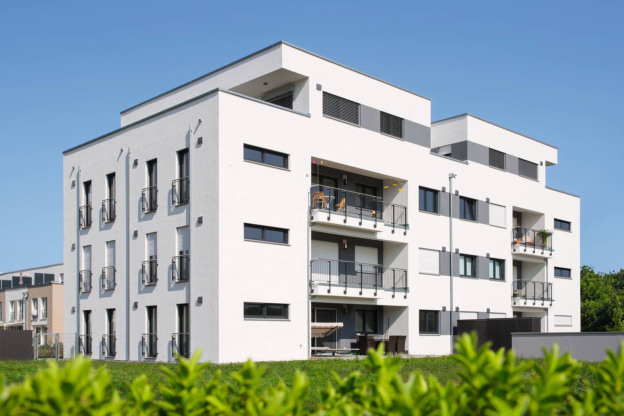Ein modernes Mehrfamilienhaus mit weißen Wänden und mehreren Balkonen. Die Fassade ist mit dunklen Fenstern und grauen Akzenten gestaltet. Im Vordergrund sind grüne Pflanzen sichtbar. Der Himmel ist klar und blau.