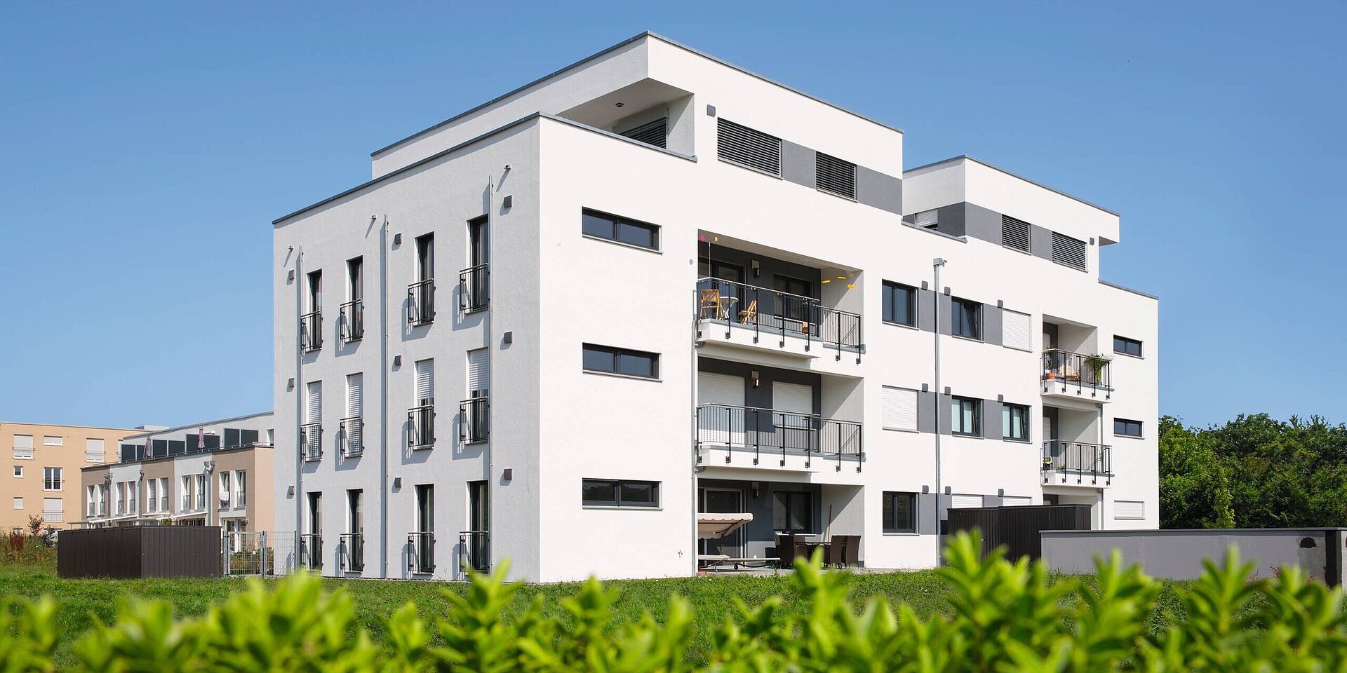 Ein modernes Mehrfamilienhaus mit weißen Wänden und mehreren Balkonen. Die Fassade ist mit dunklen Fenstern und grauen Akzenten gestaltet. Im Vordergrund sind grüne Pflanzen sichtbar. Der Himmel ist klar und blau.