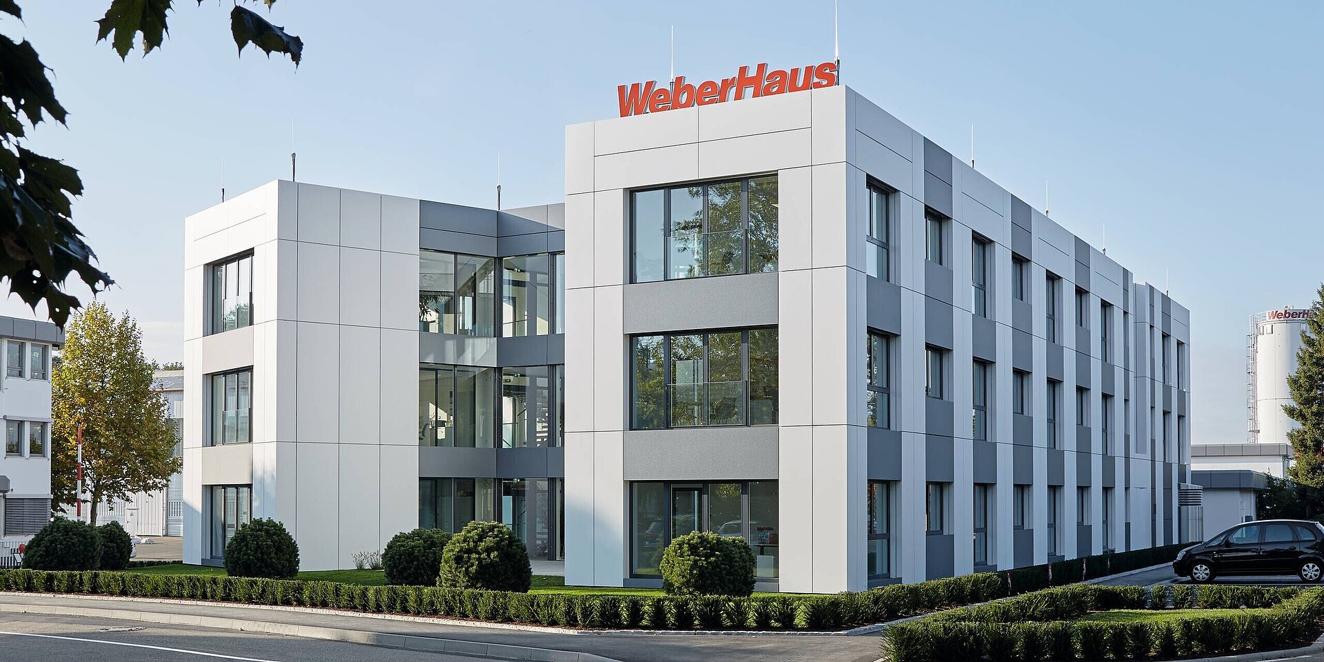 Das Bild zeigt das Verwaltungsgebäude von WeberHaus, mit klaren Linien und großen Fenstern. Die Fassade ist in Grau- und Weißtönen gestaltet. Der Unternehmensname "WeberHaus" ist auffällig in Rot über dem Eingang angebracht. 