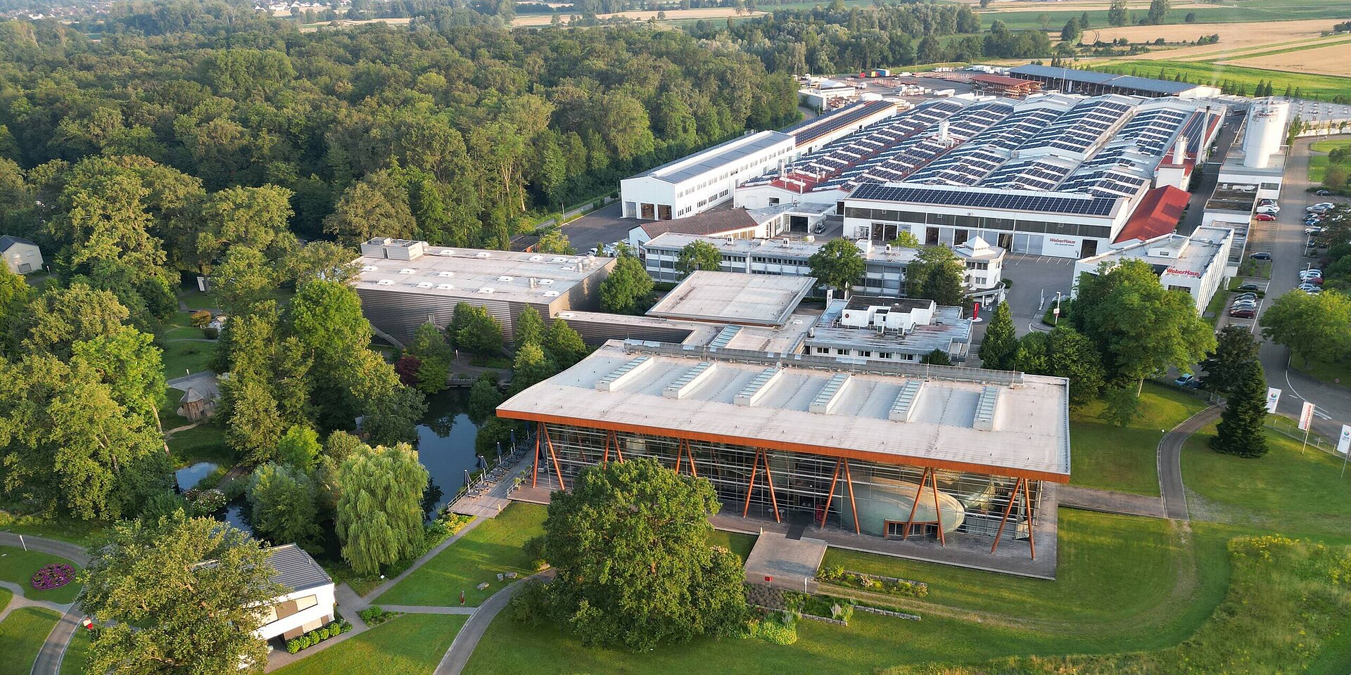 Luftaufnahme des WeberHaus Geländes, das von üppigem Grün umgeben ist. Im Vordergrund befindet sich ein modernes Gebäude mit einer großen Glasfront und Holzdetails. Im Hintergrund sind mehrere Lagerhäuser und Produktionsstätten sichtbar.