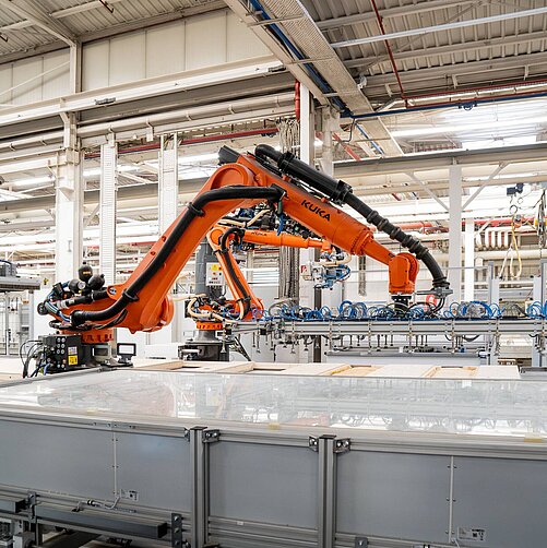 Im Bild ist ein industrieller Roboterarm in einer Fertigungsanlage zu sehen. Der Roboterführt präzise Bewegungen aus, während er an einem Förderband arbeitet. Im Hintergrund sind weitere Maschinen und Ausstattungen sichtbar, die zur Produktion beitragen.