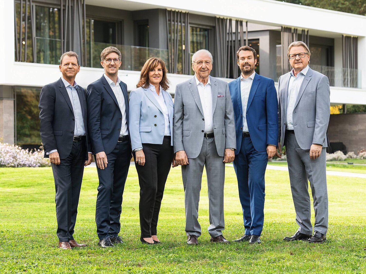 Die WeberHaus Geschäftsführung steht in formeller Kleidung auf einer Wiese vor einem modernen Gebäude. Sie lächeln und posieren für das Foto. Von links nach rechts sind Gerd Manßhard, Dr. Manuel Schönwitz, Heidi Weber-Mühleck, Hans Weber, Stephan Jager und Wolfgang Weber zu sehen.