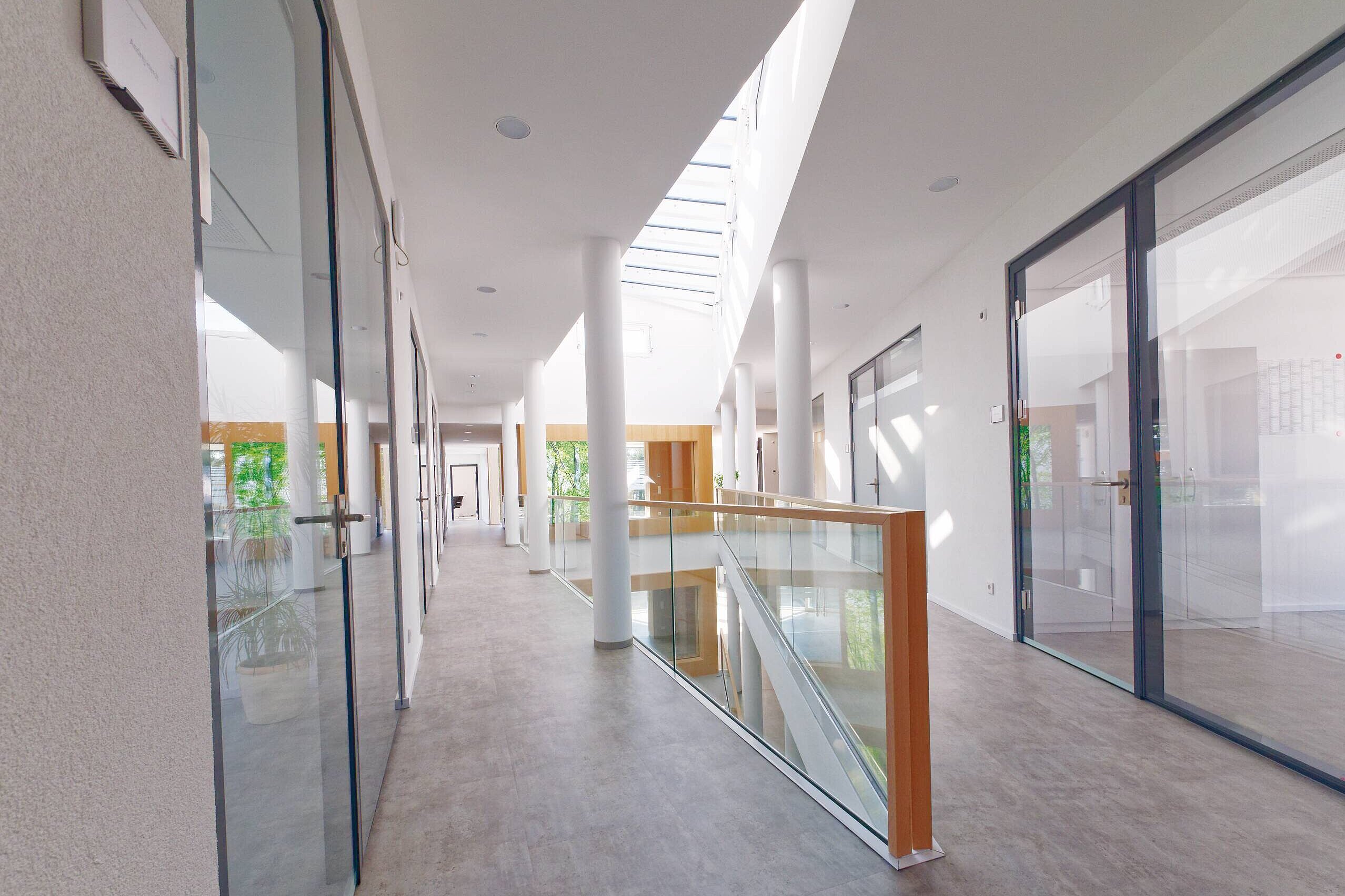 Der Bildinhalt zeigt einen modernen Flur mit großen Fenstern, die viel natürliches Licht hereinlassen. An den Wänden sind Türen zu Büros oder Räumen. Eine Geländerstruktur aus Glas und Holz verläuft entlang einer Treppe, die dem Raum eine offene und einladende Atmosphäre verleiht.