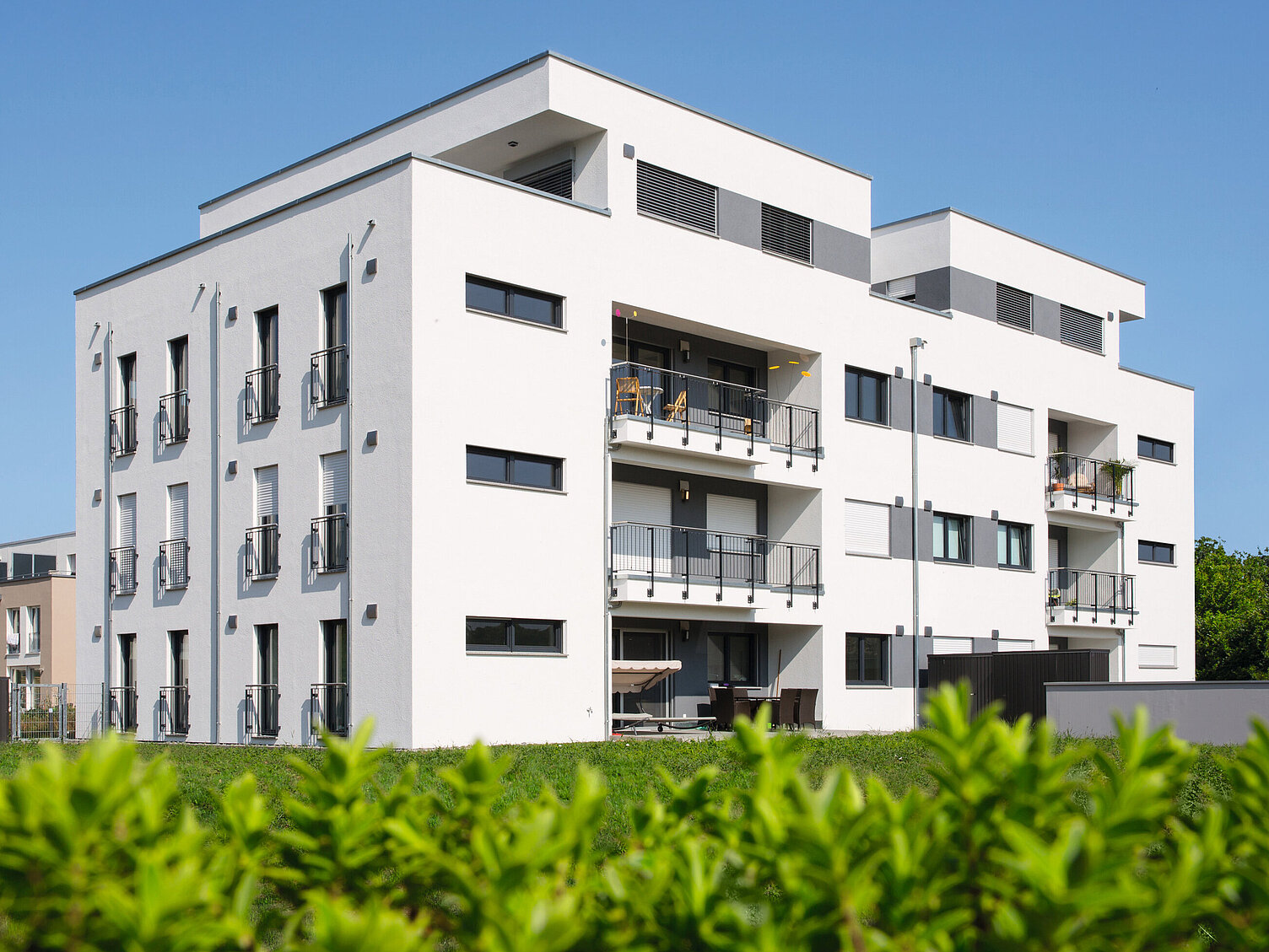 Ein modernes, mehrstöckiges Wohngebäude mit weißen Wänden und großen Fenstern. Mehrere Balkone sind sichtbar. Im Hintergrund sind weitere Gebäude und ein klarer blauer Himmel zu sehen. Die Umgebung ist von grünen Pflanzen umgeben.