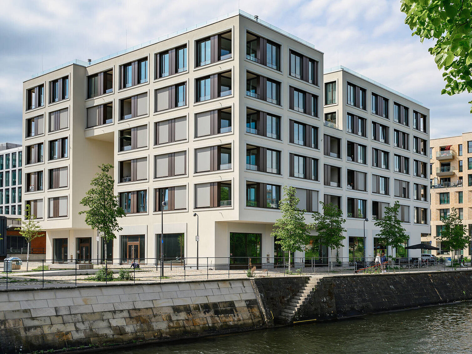 Moderne Wohngebäude am Ufer eines Flusses, umgeben von Bäumen. Die Architektur ist funktional mit großen Fenstern und klaren Linien. Im Vordergrund verläuft ein gepflasterter Weg entlang des Wassers. Das Wetter ist überwiegend bewölkt, aber es gibt Licht, das die Szene beleuchtet.