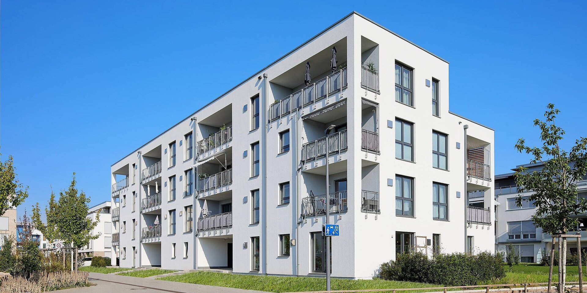 Ein modernes, weißes Mehrfamilienhaus mit mehreren Balkonen und großen Fenstern, umgeben von Grünflächen. Der Himmel ist klar und blau. Eine Rad- und Gehweg ist im Vordergrund sichtbar.
