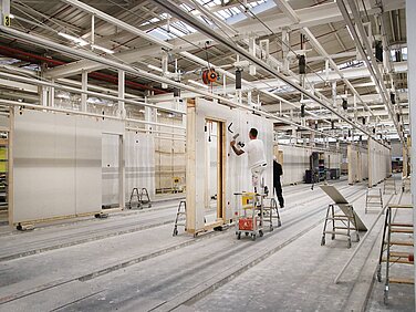 In einer großen Werkhalle arbeiten zwei Männer an der Produktion von Wänden. Ein Mitarbeiter steht auf einer Leiter und streicht eine Wand, während der andere am Boden steht. Die Umgebung ist geprägt von unvollendeten Wänden und Arbeitsgeräten.