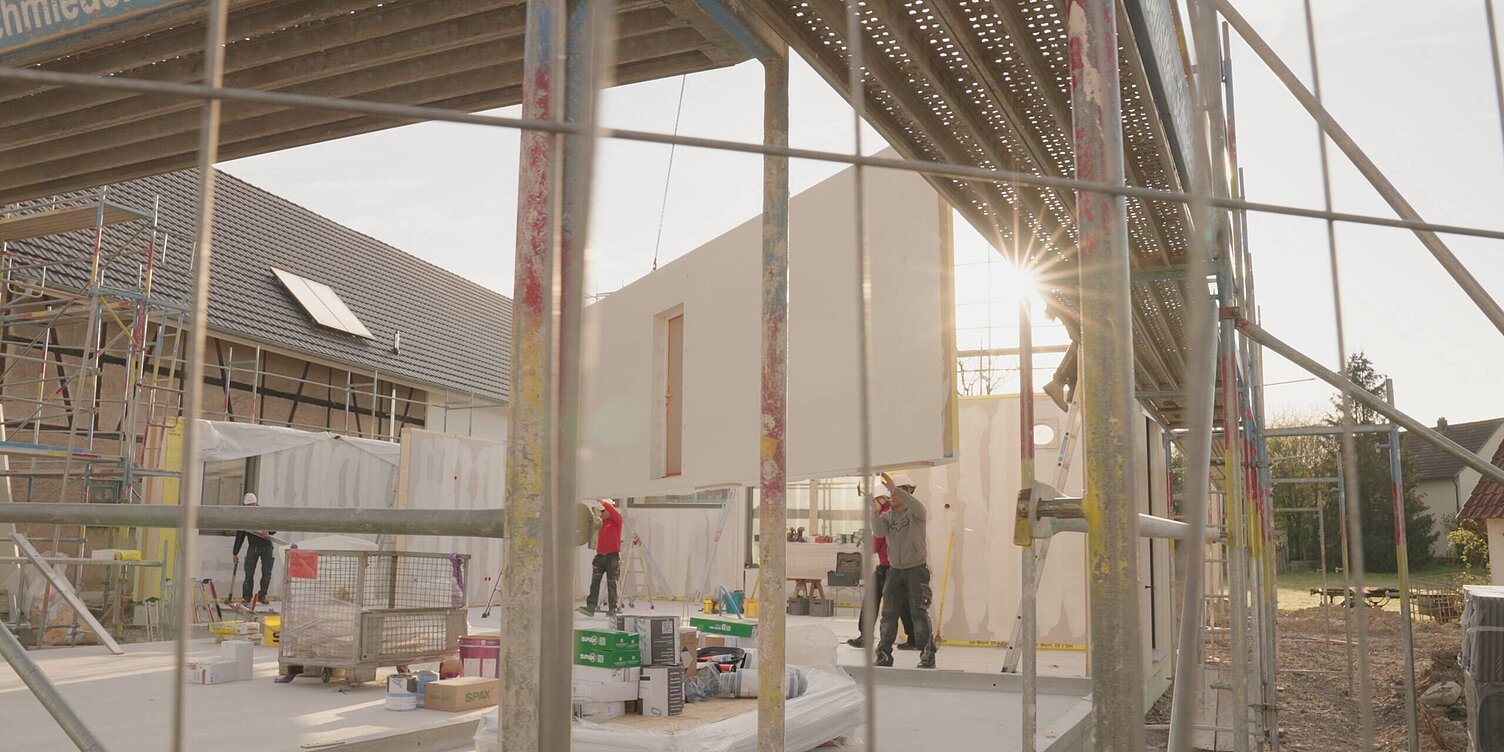 WeberHaus-Baustelle Eine WeberHaus-Baustelle mit mehreren Bauarbeitern, die Wände heben und an einem Neubau arbeiten. Gerüste und Baumaterialien sind sichtbar, während die Sonne hinter dem Gebäude scheint. Die Szene zeigt aktive Bautätigkeit und den Fortschritt des Bauvorhabens.