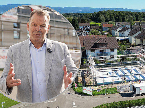 Ein Mann im Anzug steht im Vordergrund und spricht mit ausgebreiteten Händen. Im Hintergrund ist eine Baustelle mit einem Rohbau und Baugerüst sichtbar, umgeben von Wohnhäusern und einer ländlichen Landschaft.