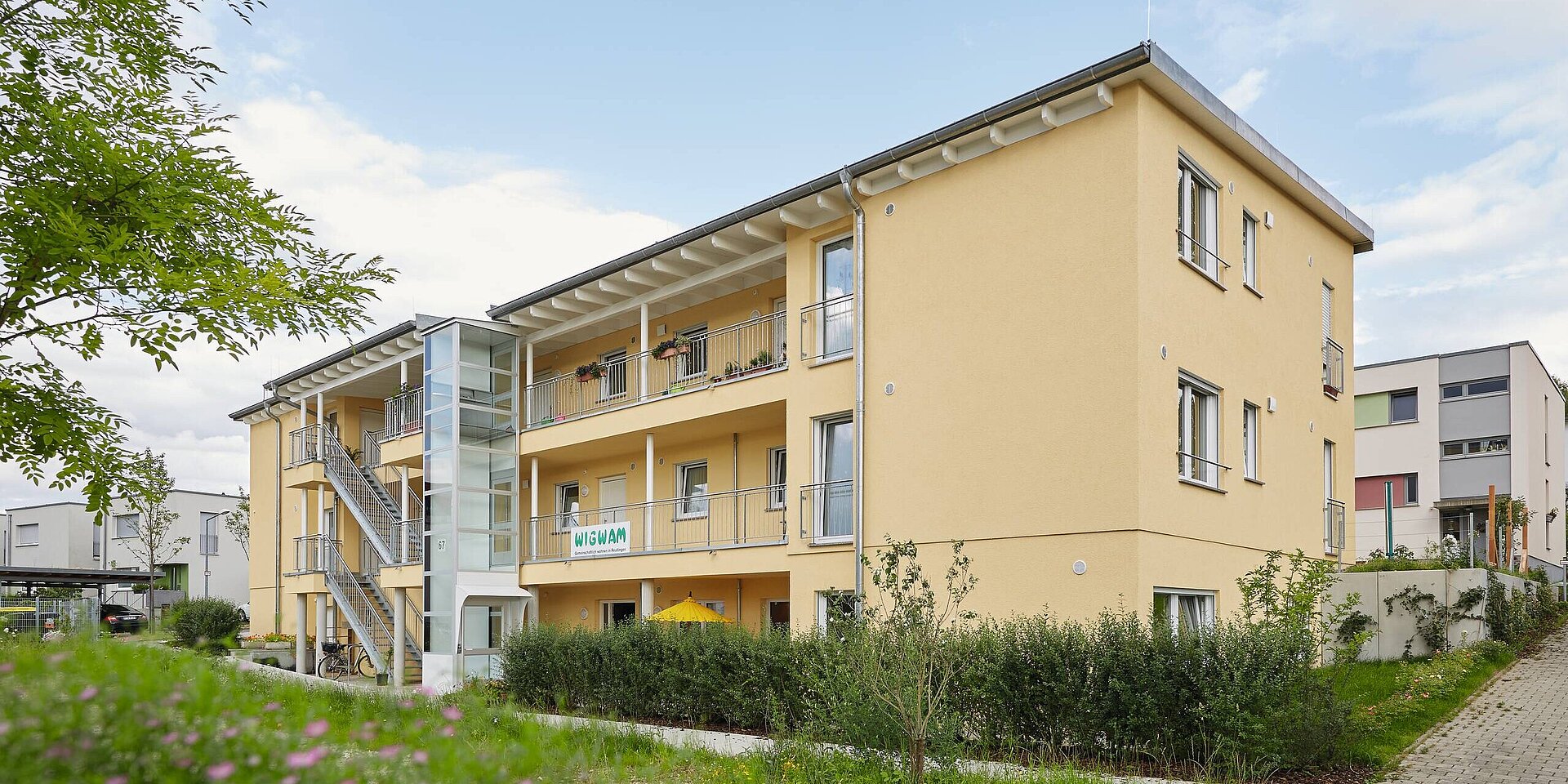 Ein modernes, gelbes Mehrfamilienhaus mit mehreren Etagen und einem gläsernen Aufzug. Die Fassade ist mit Pflanzen geschmückt, und es sind Balkone zu sehen. Im Vordergrund gibt es einen kleinen Garten und einen Weg, der zum Eingang führt.