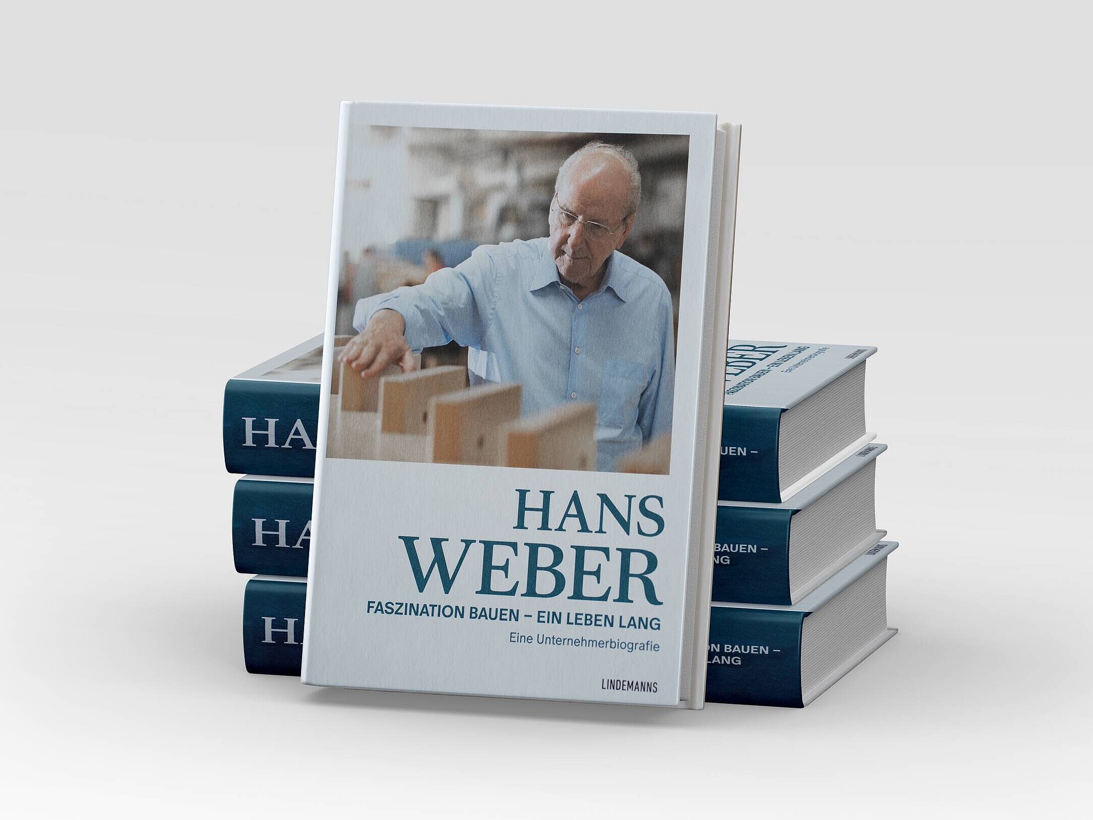 Ein Buch mit dem Titel „Hans Weber: Faszination bauen – ein Leben lang“ steht im Vordergrund. Auf dem Cover ist ein älterer Mann zu sehen, der konzentriert an einem Tisch mit Holzteilen arbeitet. Im Hintergrund sind unscharfe Werkstattutensilien zu erkennen.