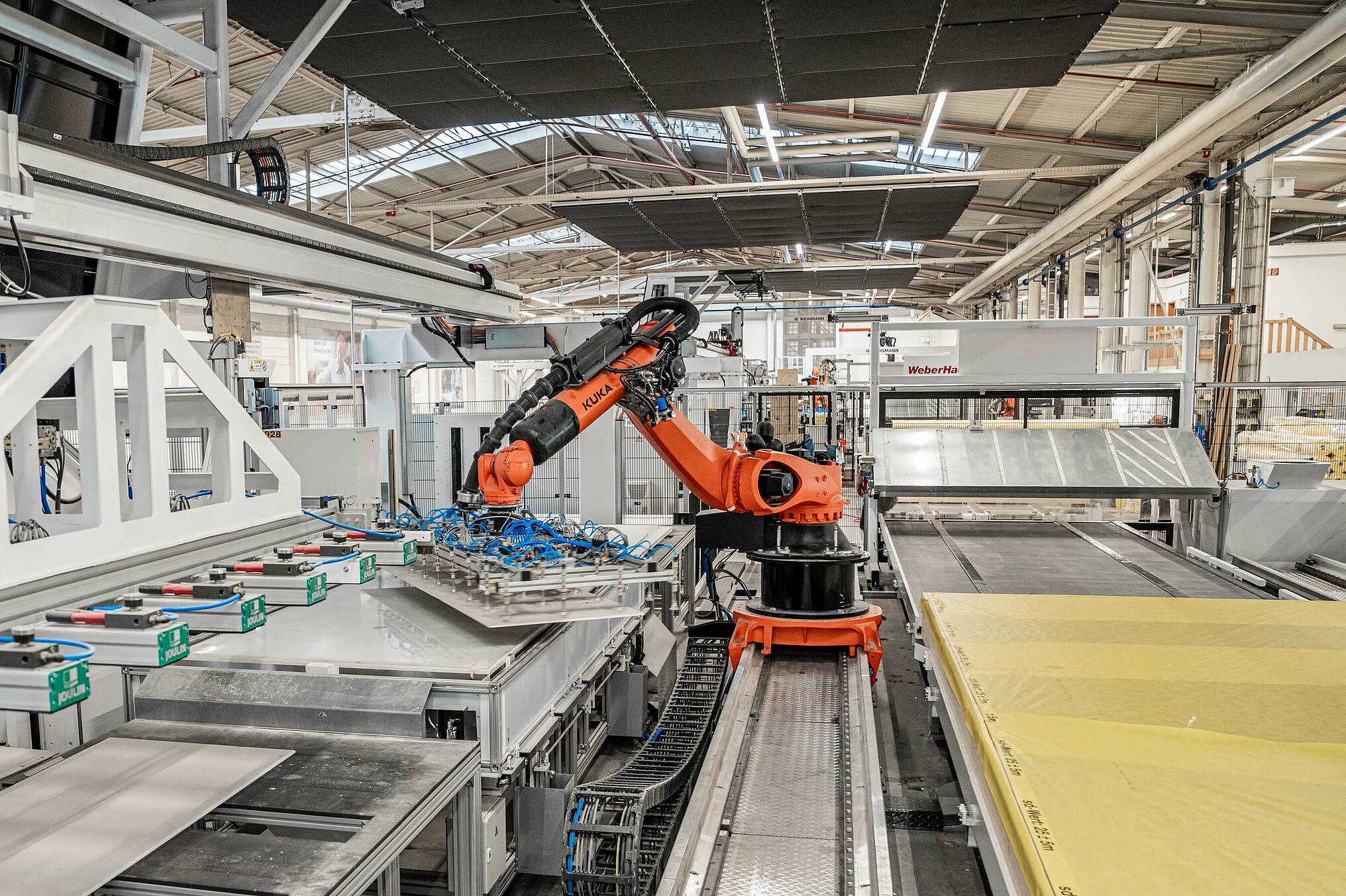 Robotik im Hausbau Ein industrieller Roboterarm steht in einer modernen Fertigungsanlage. Der Roboter greift eine Platte von einem Förderband, während im Hintergrund verschiedene Maschinen und Materialien sichtbar sind. Die Umgebung ist gut beleuchtet und zeigt eine effiziente Produktionsumgebung.