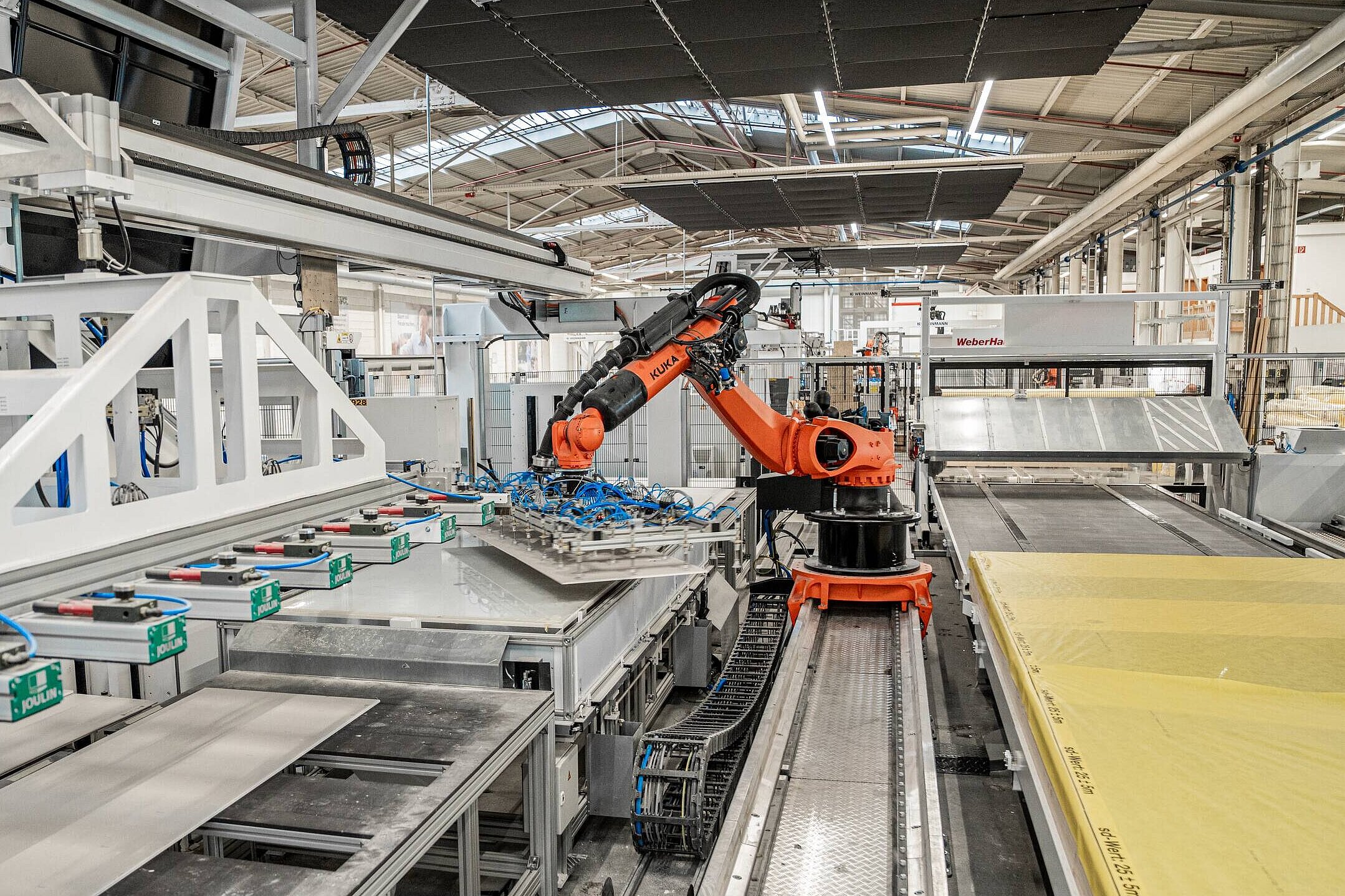 Robotik im Hausbau Ein industrieller Roboterarm steht in einer modernen Fertigungsanlage. Der Roboter greift eine Platte von einem Förderband, während im Hintergrund verschiedene Maschinen und Materialien sichtbar sind. Die Umgebung ist gut beleuchtet und zeigt eine effiziente Produktionsumgebung.