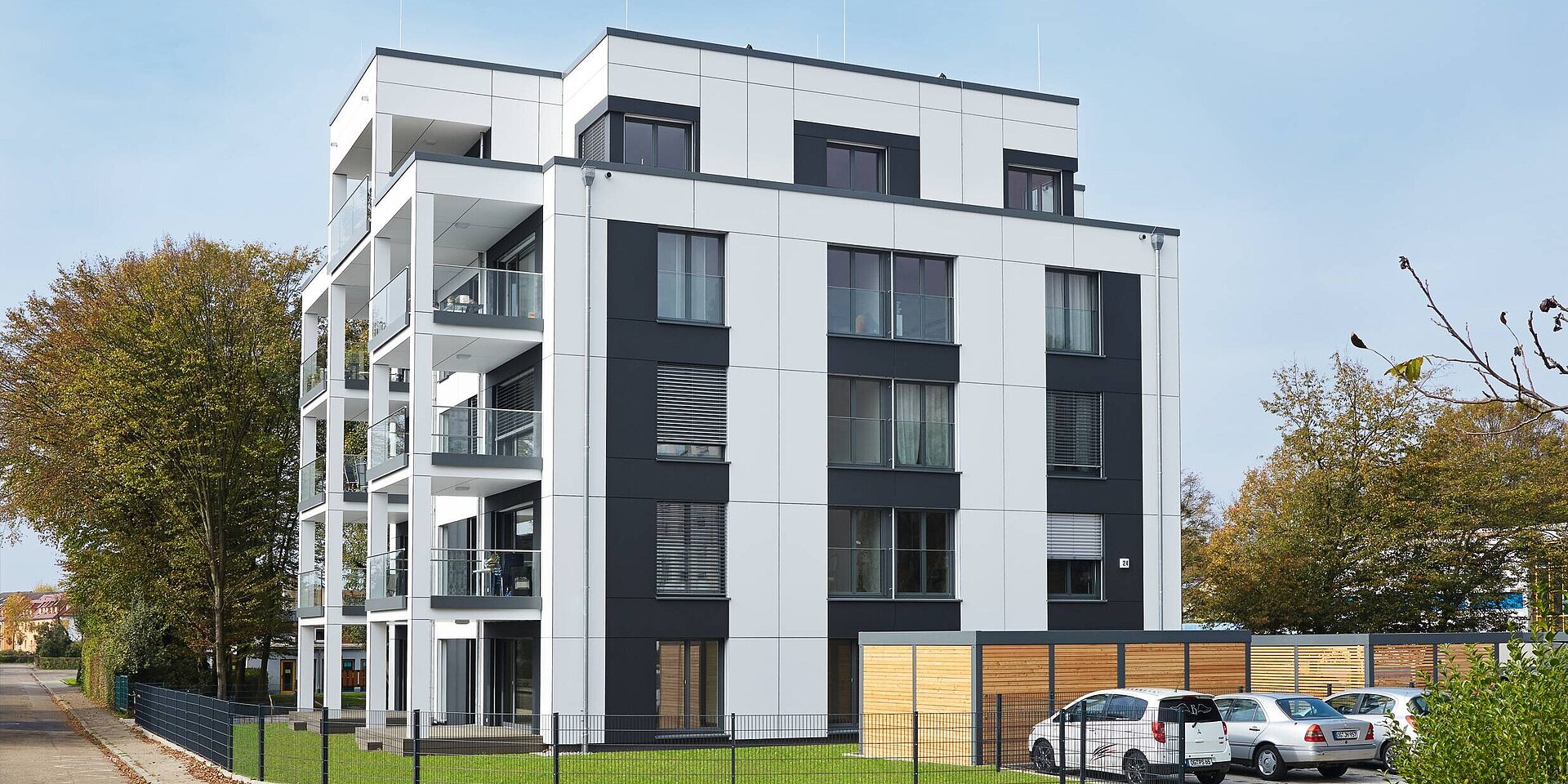 Das Bild zeigt ein modernes, Mehrfamilienhaus in einem städtischen Umfeld. Die Fassade ist in Schwarz und Weiß gehalten, mit großen Fenstern und Balkonen. Vor dem Gebäude befindet sich ein gepflegter Rasen und ein parkähnlicher Bereich mit Bäumen sowie Parkplätze.